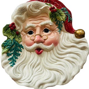 May include: A ceramic Santa Claus face with a white beard and mustache, rosy cheeks, and a red hat. The Santa figure has gold-rimmed glasses and is decorated with holly and berries. The overall design is festive and detailed.