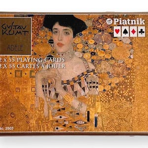 Könnte beinhalten: Ein Kartenspiel-Set in einer Schachtel mit Gustav Klimts Porträt von Adele Bloch-Bauer. Die Schachtel hat ein Gold- und Braunschema mit dem Text "GUSTAV KLIMT ADELE" und "2 x 55 SPIELKARTEN". Das Piatnik-Logo ist sichtbar.