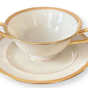 Könnte beinhalten: Cremefarbene Teetasse mit zwei Henkeln und passender Untertasse. Beide Teile haben einen goldenen Rand. Die Teetasse und die Untertasse sind vor einem weißen Hintergrund platziert.