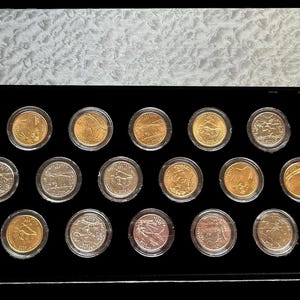 Pode incluir: Uma coleção de vinte moedas exibidas numa caixa de apresentação preta. As moedas são de várias cores, incluindo ouro, prata e cobre. A caixa tem uma tampa texturizada de cor clara.
