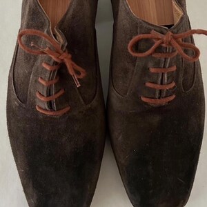 May include: A pair of brown suede dress shoes with orange laces. The shoes have a classic Oxford style with a square toe. The suede material has a soft, textured appearance. The laces are neatly tied, and the shoes appear unworn.