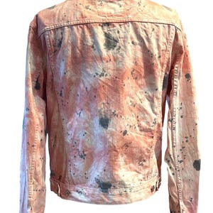 Op de afbeelding: Een roze denim jack met een splatter paint design in grijstinten en wit.