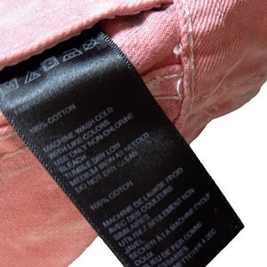 Op de afbeelding: Een zwart waslabel met witte tekst met wasinstructies voor een kledingstuk van 100% katoen. Het label luidt "Machinewas koud met soortgelijke kleuren, alleen niet-chloorbleekmiddel gebruiken, drogen in de droger op lage temperatuur, strijken op middelhoge temperatuur indien nodig, niet chemisch reinigen".