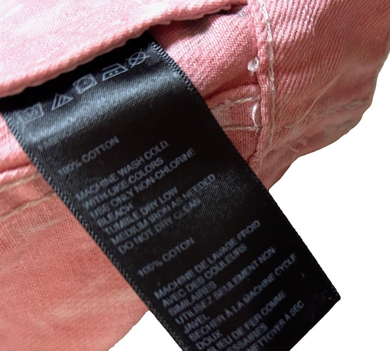 Op de afbeelding: Een zwart waslabel met witte tekst met wasinstructies voor een kledingstuk van 100% katoen. Het label luidt "Machinewas koud met soortgelijke kleuren, alleen niet-chloorbleekmiddel gebruiken, drogen in de droger op lage temperatuur, strijken op middelhoge temperatuur indien nodig, niet chemisch reinigen".