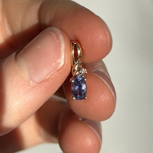 Pode incluir: Um pingente de ouro com uma pequena pedra preciosa oval roxa e um pequeno detalhe de diamante. O pingente &eacute; segurado entre os dedos contra um fundo branco. A configura&ccedil;&atilde;o de ouro tem um design torcido.