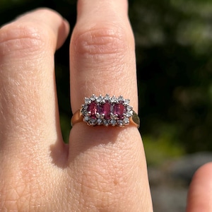 May include: A gold ring with three rectangular, deep red gemstones, encircled by smaller, clear stones. The ring is set against a blurred green backdrop, highlighting the jewellery's design and colour.