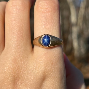 Pode incluir: Um anel de ouro com uma pedra preciosa de safira estrela azul. O anel tem uma banda simples e uma moldura que segura a pedra preciosa oval. A safira estrela tem um padrão de estrela de seis raios. O anel é mostrado em uma mão.
