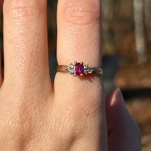 May include: A gold ring with a central, rectangular, deep red gemstone, with two smaller clear stones on either side. The ring is set against a blurred outdoor background, highlighting the jewellery's design and colour.