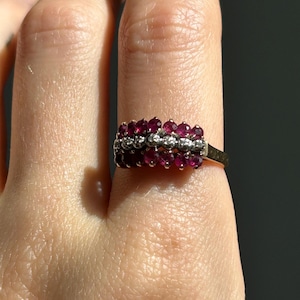 May include: A gold ring featuring three horizontal rows of stones. The top and bottom rows consist of small, round, deep red rubies, while the centre row is composed of smaller, clear, round diamonds set in a bright, polished metal band.