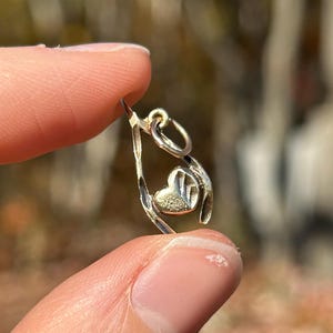May include: A small, silver-colored charm with a heart-shaped design. The charm is held between two fingers against a blurred background of fall foliage. The charm has a loop at the top for attaching to a necklace or bracelet.