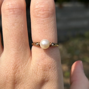 Pode incluir: Um anel de ouro com uma pérola branca central e dois pequenos diamantes. O anel é colocado em uma fina banda de ouro, mostrando um design clássico e elegante. O anel é mostrado em um dedo.