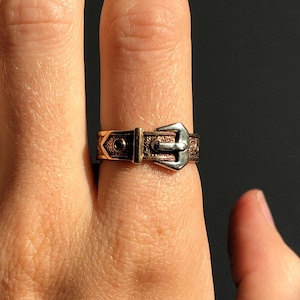 Pode incluir: Um anel em tom bronze com um design de fivela. O anel apresenta um fecho de fivela detalhado e elementos decorativos ao longo da banda. O anel é usado num dedo, mostrando o intrincado trabalho em metal e a estética vintage.