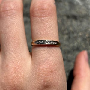 Peut inclure: Une bague en or avec une rangée de petits diamants transparents. La bague est portée au doigt, la main étant au premier plan. Le design est simple et élégant, idéal pour un usage quotidien ou des occasions spéciales.