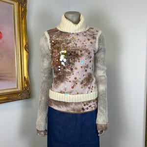 May include: A turtleneck sweater with a cream-colored collar and cuffs. The bodice features a pink and brown pattern with sequins. The sleeves are a light gray knit, and the skirt is a dark blue denim.