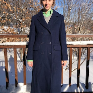 May include: A long, navy blue double-breasted coat with a high collar. The coat is being worn by a person standing in front of a wooden railing with snow on the ground.