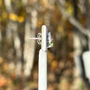 K&ouml;nnte beinhalten: Ein silberner Ohrstecker mit einem kleinen, runden, hellgr&uuml;nen Edelstein und einigen kleineren, klaren Edelsteinen. Der Ohrring befindet sich auf einer wei&szlig;en Pr&auml;sentationskarte mit verschwommenem Herbstlaub im Hintergrund.
