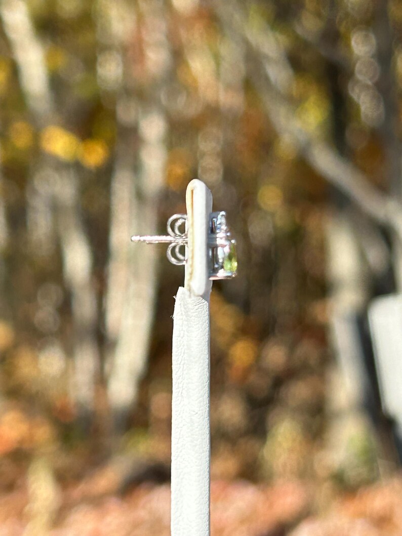 K&ouml;nnte beinhalten: Ein silberner Ohrstecker mit einem kleinen, runden, hellgr&uuml;nen Edelstein und einigen kleineren, klaren Edelsteinen. Der Ohrring befindet sich auf einer wei&szlig;en Pr&auml;sentationskarte mit verschwommenem Herbstlaub im Hintergrund.