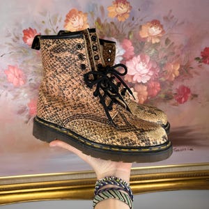 Könnte beinhalten: Ein Paar braune und beige Combat-Stiefel mit Schlangenmuster, schwarzen Schnürsenkeln und einer schwarzen Sohle. Die Stiefel haben ein klassisches Schnürdesign mit mehreren Ösen. Die Stiefel werden von einer Hand gehalten.