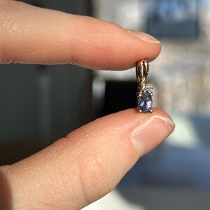 Pode incluir: Um pingente de ouro com uma pequena pedra preciosa oval roxa e um pequeno detalhe de diamante. O pingente &eacute; segurado entre dois dedos, mostrando seu design delicado e artesanato.