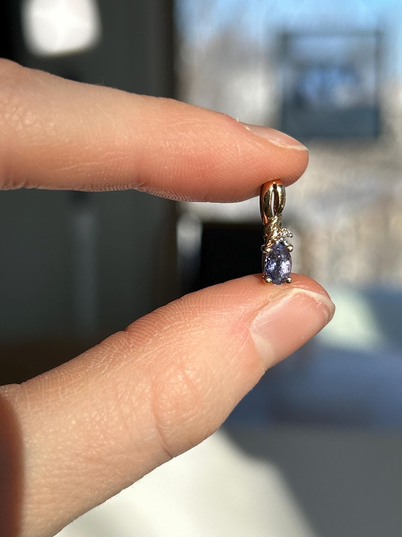 Pode incluir: Um pingente de ouro com uma pequena pedra preciosa oval roxa e um pequeno detalhe de diamante. O pingente &eacute; segurado entre dois dedos, mostrando seu design delicado e artesanato.