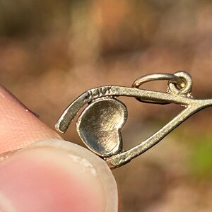 May include: Close-up of a gold-colored pendant with a heart-shaped charm. The pendant has a delicate design with a small loop at the top. The text "14K" is visible on the pendant. The background is blurred.