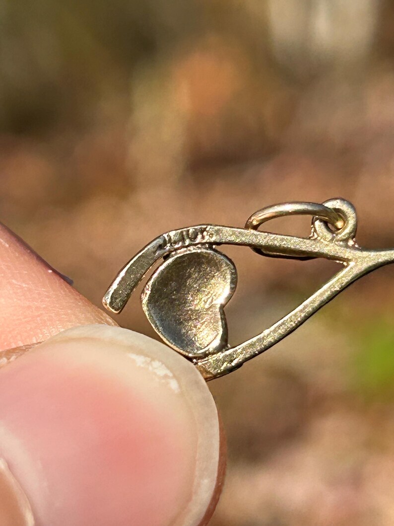 May include: Close-up of a gold-colored pendant with a heart-shaped charm. The pendant has a delicate design with a small loop at the top. The text "14K" is visible on the pendant. The background is blurred.