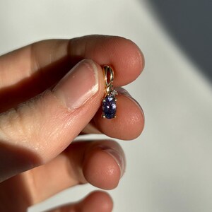 Pode incluir: Um pequeno pingente de ouro com uma pedra preciosa oval roxa e uma pequena pedra preciosa branca. O pingente &eacute; segurado entre os dedos contra um fundo branco. A configura&ccedil;&atilde;o de ouro tem um pequeno la&ccedil;o na parte superior.
