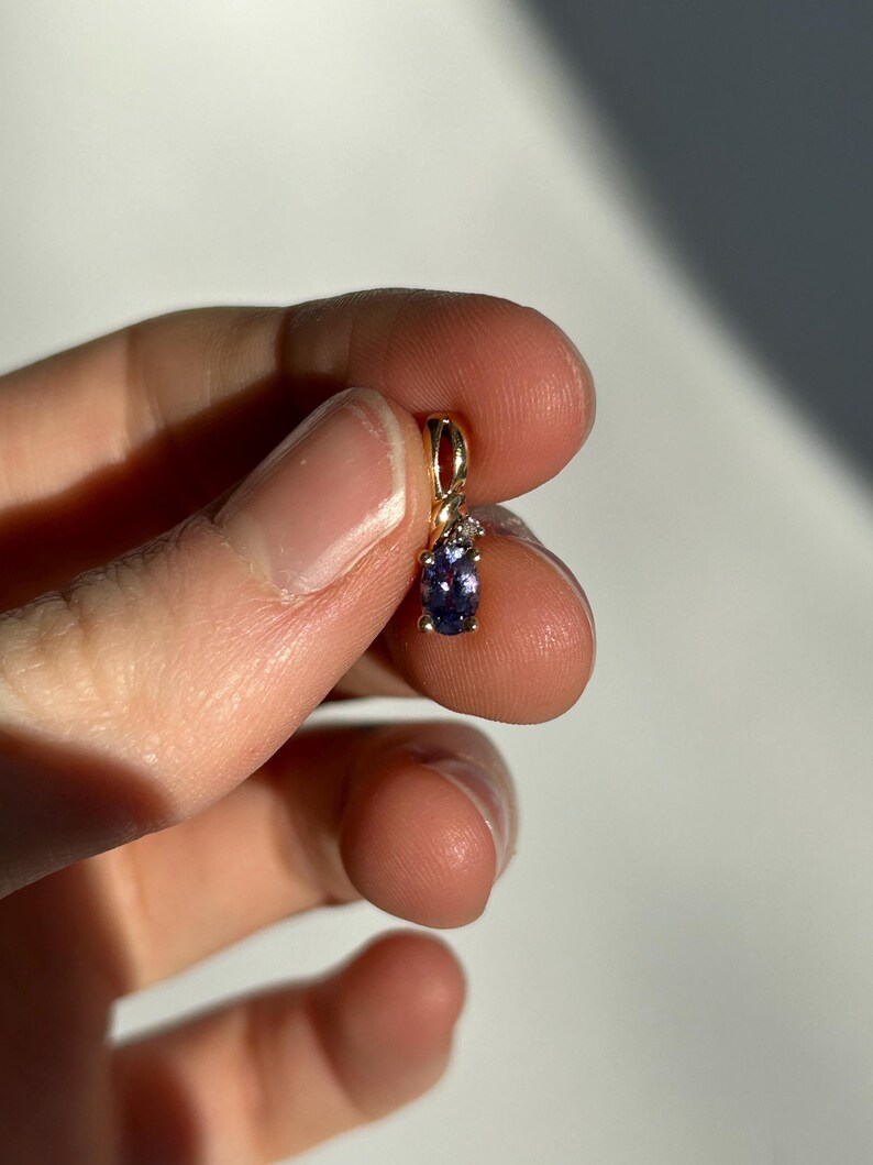 Pode incluir: Um pequeno pingente de ouro com uma pedra preciosa oval roxa e uma pequena pedra preciosa branca. O pingente &eacute; segurado entre os dedos contra um fundo branco. A configura&ccedil;&atilde;o de ouro tem um pequeno la&ccedil;o na parte superior.