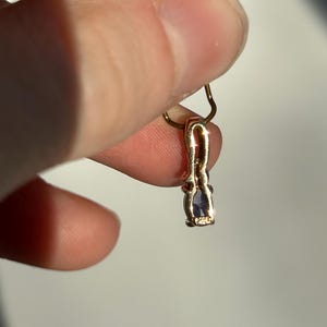 Pode incluir: Um pingente de ouro com uma pequena pedra preciosa roxa em forma oval. O pingente &eacute; preso por uma corrente de ouro. O pingente &eacute; segurado entre dois dedos.
