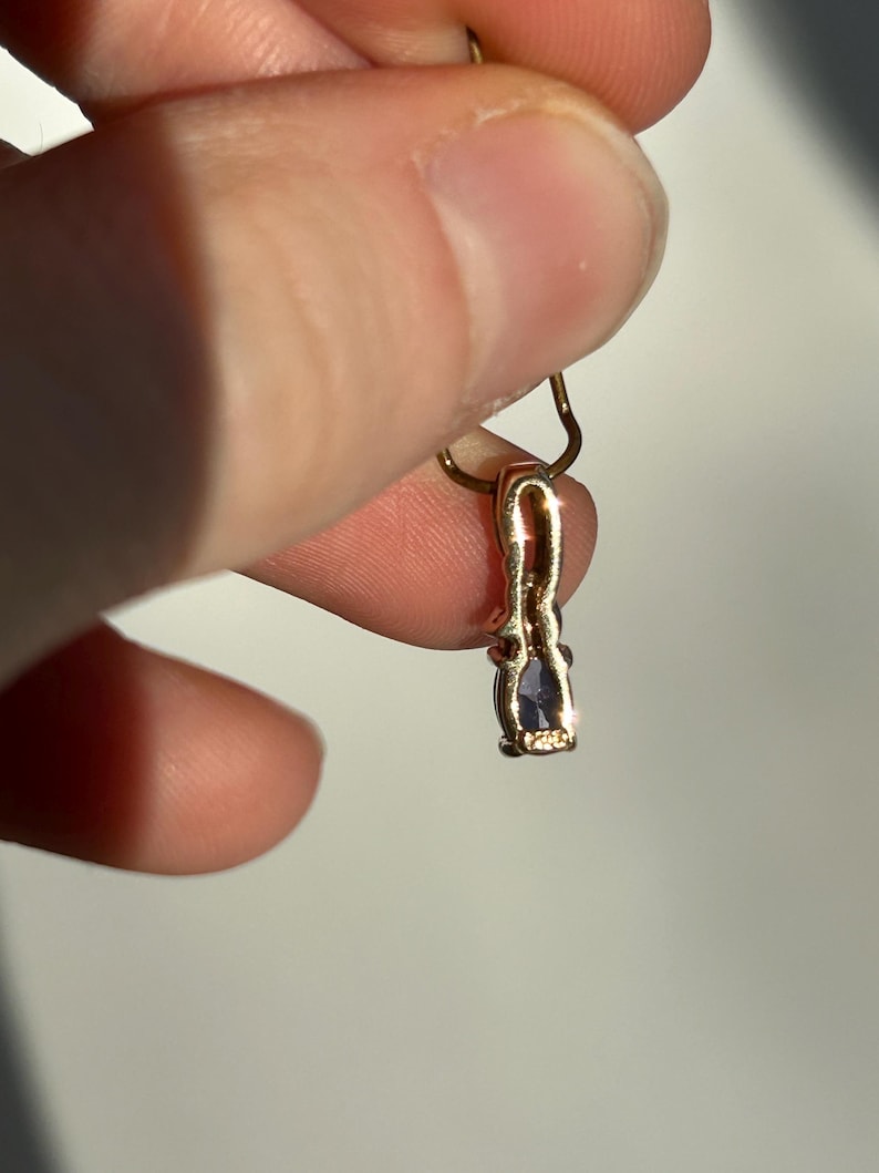 Pode incluir: Um pingente de ouro com uma pequena pedra preciosa roxa em forma oval. O pingente &eacute; preso por uma corrente de ouro. O pingente &eacute; segurado entre dois dedos.
