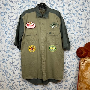 May include: A khaki button-down shirt with short sleeves and patches. The shirt has a green yoke and sleeves. Patches include "Mann's Bait Company", "Let's Talk Fishing", and other fishing-related logos. The shirt is hanging on a black hanger.