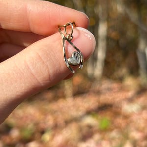 May include: A gold-colored pendant with a wishbone design, holding a small heart-shaped charm. The heart has a textured surface. The pendant is suspended from a small loop.