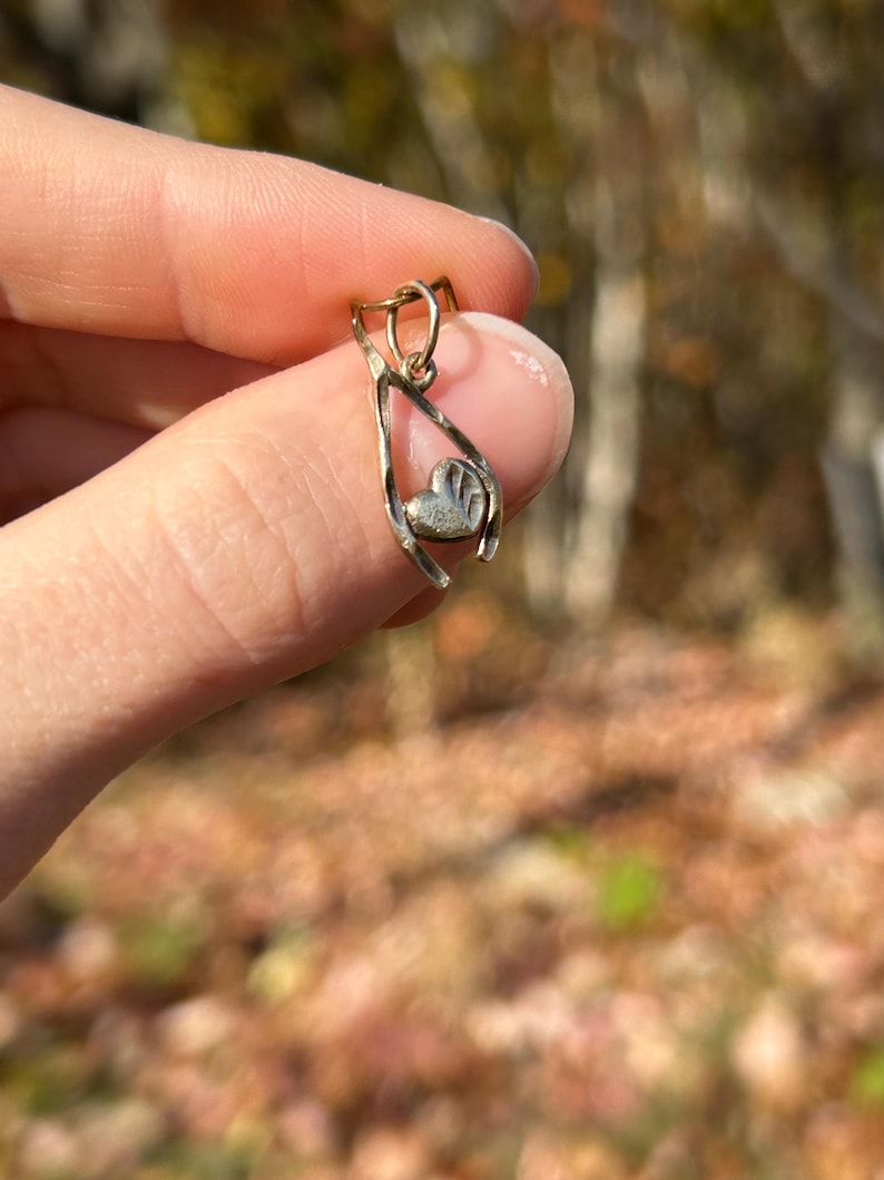 May include: A gold-colored pendant with a wishbone design, holding a small heart-shaped charm. The heart has a textured surface. The pendant is suspended from a small loop.
