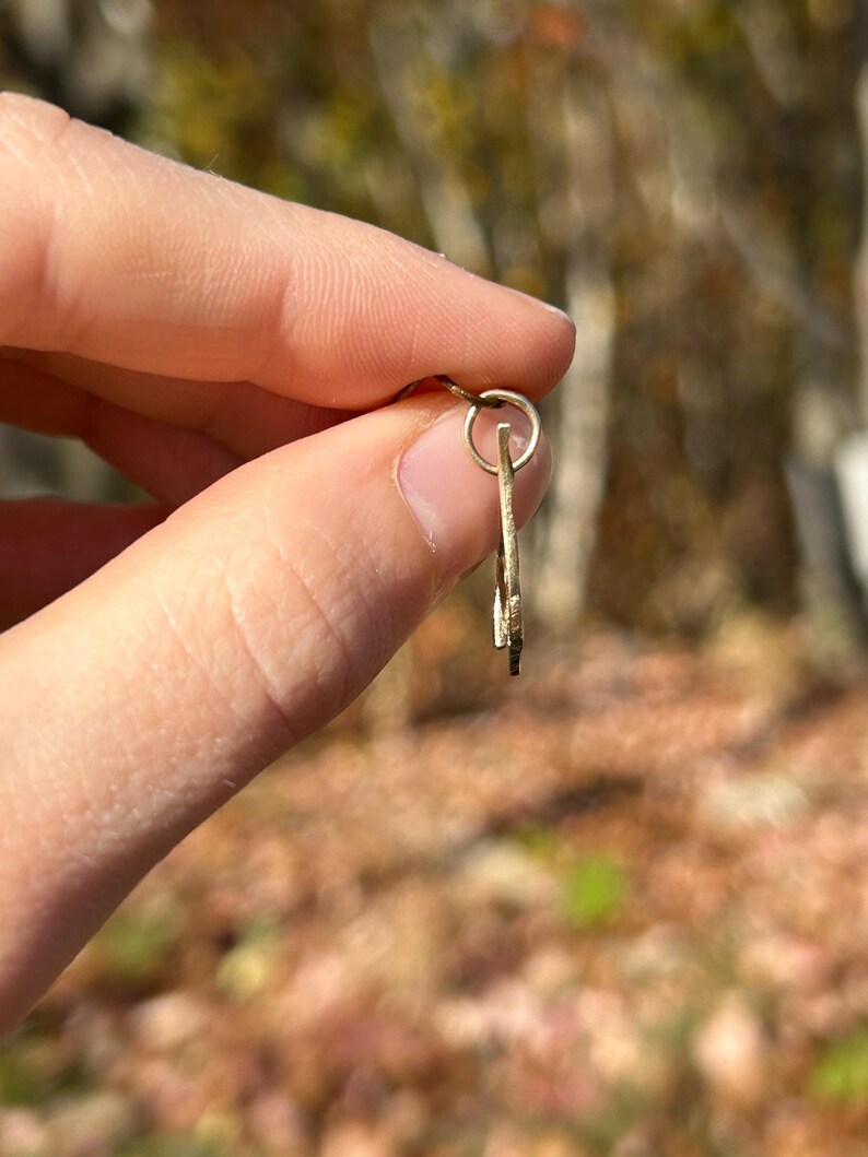 May include: A small, gold-colored key pendant with a circular loop at the top. The key has a simple, elegant design. The background is blurred, with natural colors of brown and green.