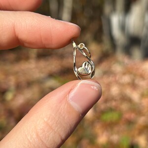 May include: A small, gold-colored pendant with a heart-shaped design. The pendant features a teardrop-shaped frame with a textured heart in the center. The jewelry is held between fingers against a blurred natural background.