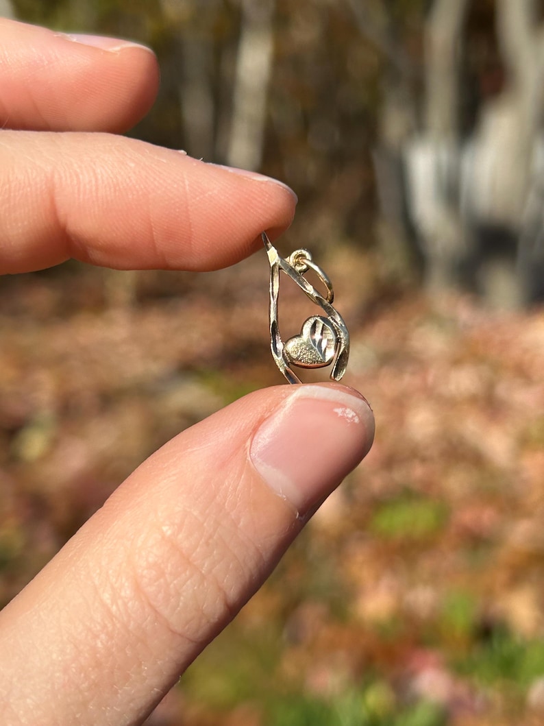 May include: A small, gold-colored pendant with a heart-shaped design. The pendant features a teardrop-shaped frame with a textured heart in the center. The jewelry is held between fingers against a blurred natural background.