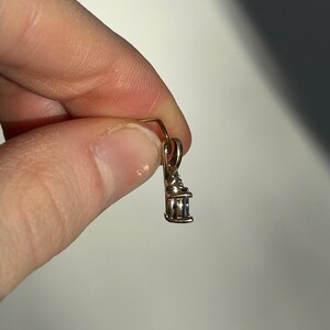 Pode incluir: Um pequeno pingente dourado com uma pedra retangular de cor clara. O pingente &eacute; segurado entre os dedos contra um fundo neutro. O design do pingente apresenta um pequeno la&ccedil;o para uma corrente.