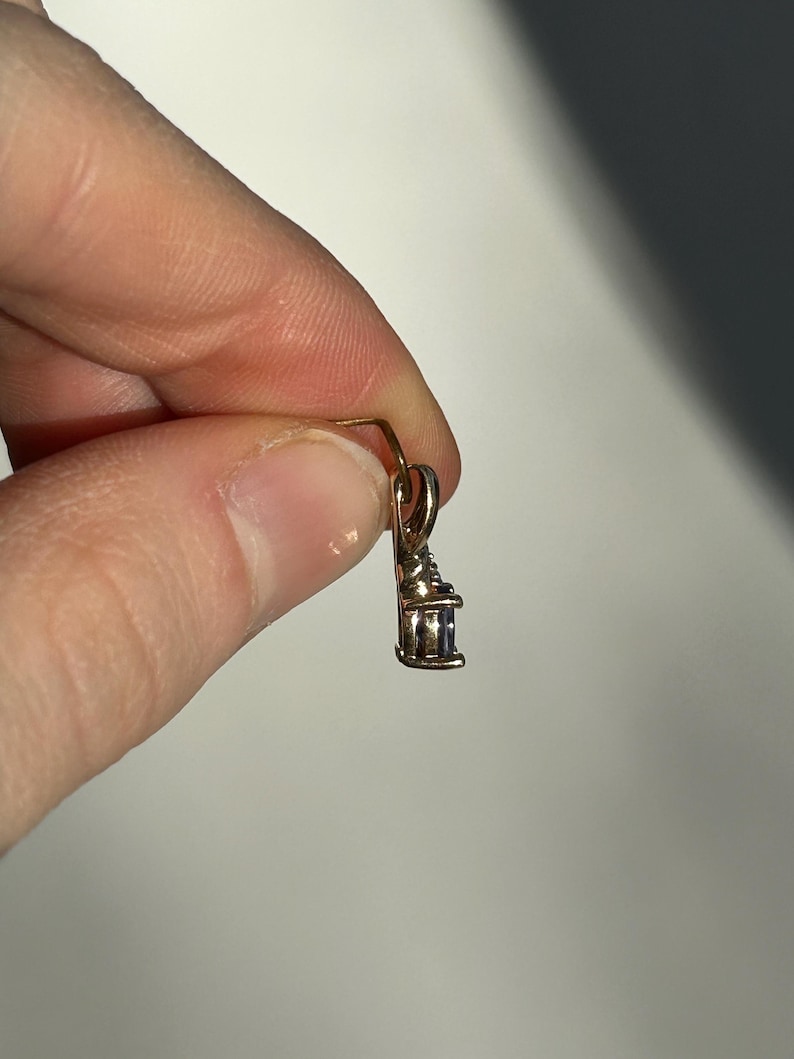 Pode incluir: Um pequeno pingente dourado com uma pedra retangular de cor clara. O pingente &eacute; segurado entre os dedos contra um fundo neutro. O design do pingente apresenta um pequeno la&ccedil;o para uma corrente.