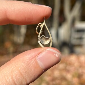 May include: A gold-colored pendant with a teardrop shape, featuring a heart-shaped design in the center. The pendant is held between fingers against a blurred outdoor background.