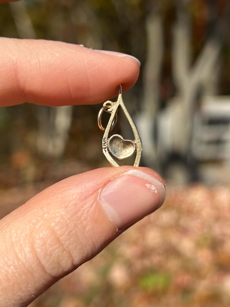 May include: A gold-colored pendant with a teardrop shape, featuring a heart-shaped design in the center. The pendant is held between fingers against a blurred outdoor background.