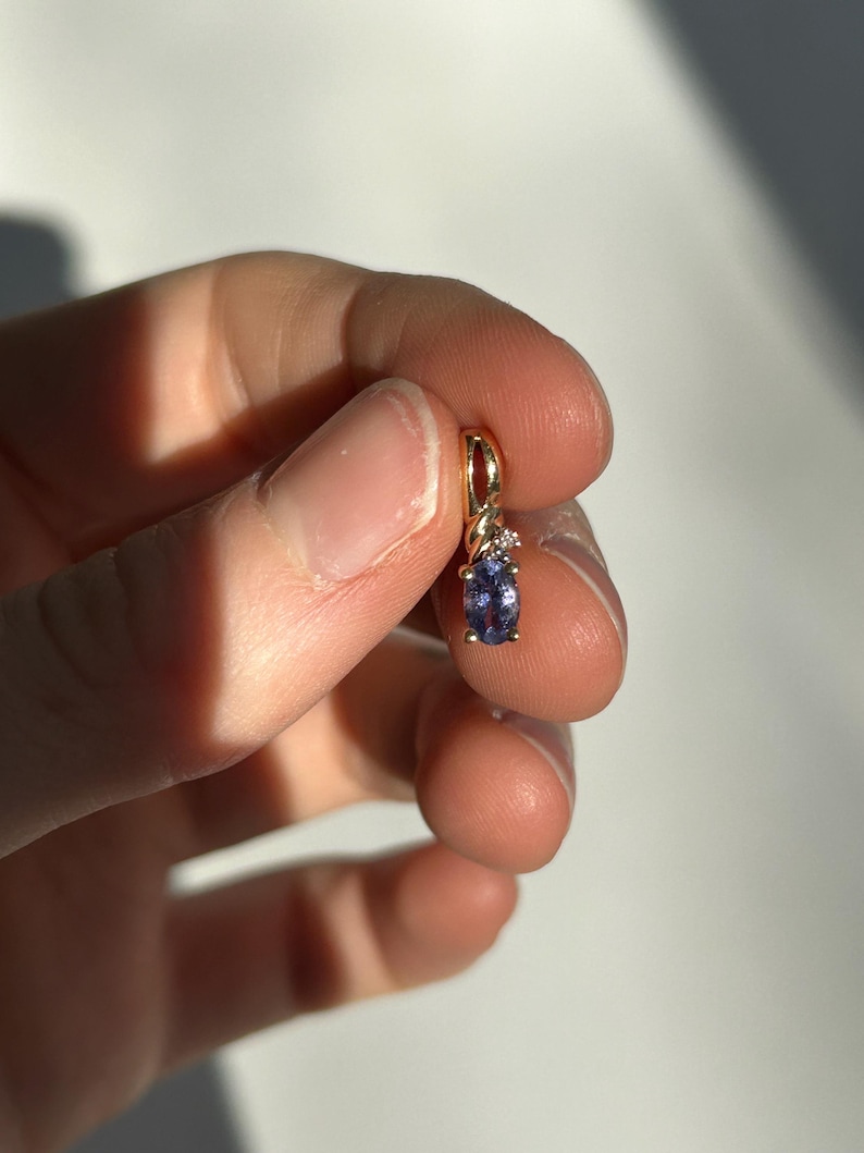 Pode incluir: Um pingente de ouro com uma pequena pedra preciosa oval roxa e um pequeno detalhe de diamante. O pingente &eacute; segurado entre os dedos contra um fundo branco. A configura&ccedil;&atilde;o de ouro tem um design torcido.