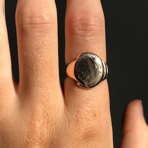 Pode incluir: Um anel de sinete dourado com uma face oval prateada polida. O anel é usado num dedo, com a banda e a face visíveis contra um fundo escuro. O design do anel é simples e clássico, adequado para uso diário.
