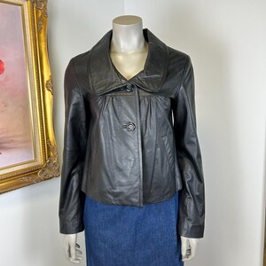 Könnte beinhalten: Schwarze Lederjacke mit breitem Kragen und Zwei-Knopf-Verschluss. Die Jacke wird auf einer Schaufensterpuppe mit einem blauen Jeansrock präsentiert. Die Jacke hat ein klassisches, maßgeschneidertes Design.
