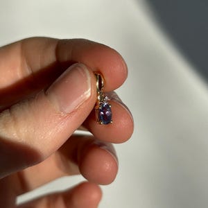 Pode incluir: Um pendente de ouro com uma pedra preciosa oval roxa escura. O pendente tem um pequeno detalhe de diamante e &eacute; suspenso por um la&ccedil;o de ouro. A joia &eacute; segurada em uma m&atilde;o contra um fundo branco.
