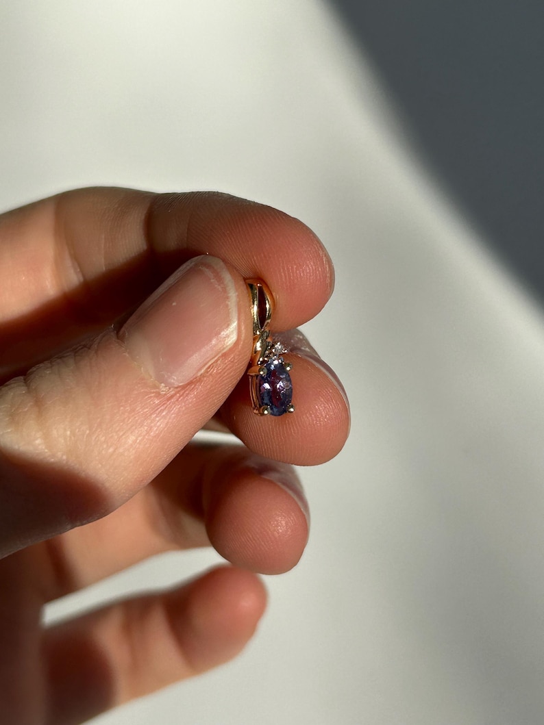 Pode incluir: Um pendente de ouro com uma pedra preciosa oval roxa escura. O pendente tem um pequeno detalhe de diamante e &eacute; suspenso por um la&ccedil;o de ouro. A joia &eacute; segurada em uma m&atilde;o contra um fundo branco.