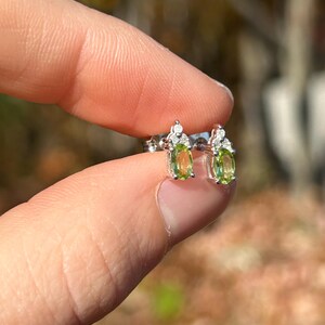 K&ouml;nnte beinhalten: Ohrstecker mit ovalen, hellgr&uuml;nen Edelsteinen, vermutlich Peridot, in Silber oder wei&szlig;em Metall gefasst. Jeder Ohrring hat kleine, klare Edelsteine, die f&uuml;r Glanz sorgen. Die Ohrringe werden zwischen Fingern vor einem unscharfen Hintergrund gehalten.