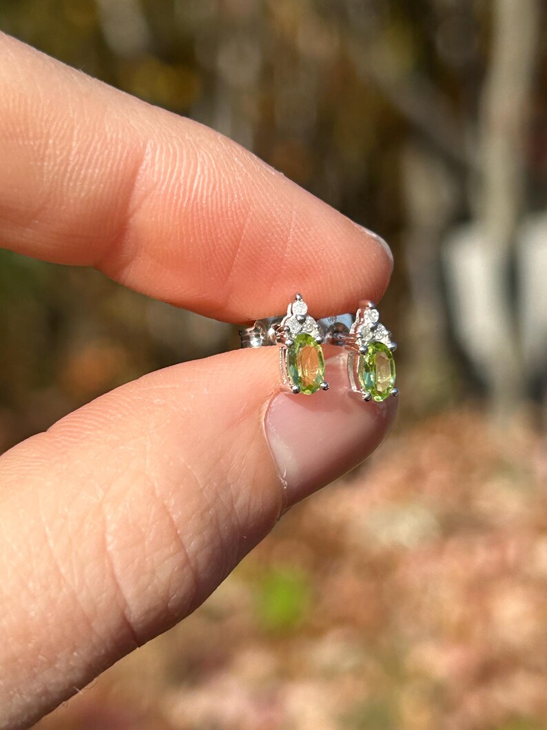 K&ouml;nnte beinhalten: Ohrstecker mit ovalen, hellgr&uuml;nen Edelsteinen, vermutlich Peridot, in Silber oder wei&szlig;em Metall gefasst. Jeder Ohrring hat kleine, klare Edelsteine, die f&uuml;r Glanz sorgen. Die Ohrringe werden zwischen Fingern vor einem unscharfen Hintergrund gehalten.