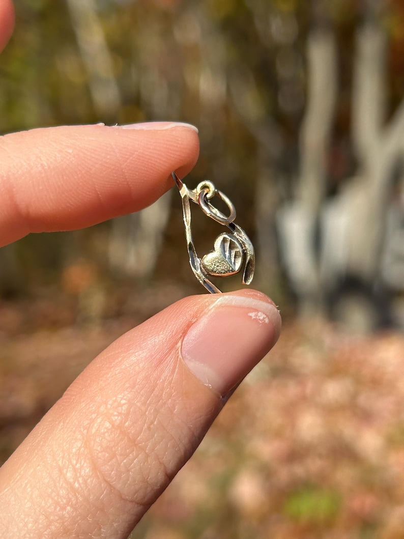 May include: A small, silver-colored charm with a heart-shaped design. The charm is held between two fingers against a blurred background of fall foliage. The charm has a loop at the top for attaching to a necklace or bracelet.
