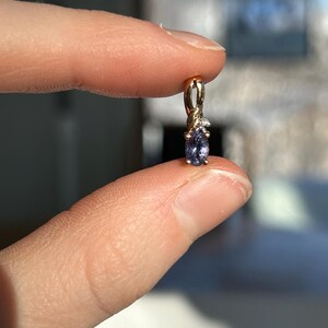 Pode incluir: Um pingente de ouro com uma pequena pedra preciosa azul em forma oval. O pingente tem um design delicado com pequenas pedras de destaque transparentes. A joia &eacute; segurada entre dois dedos, com um fundo desfocado.