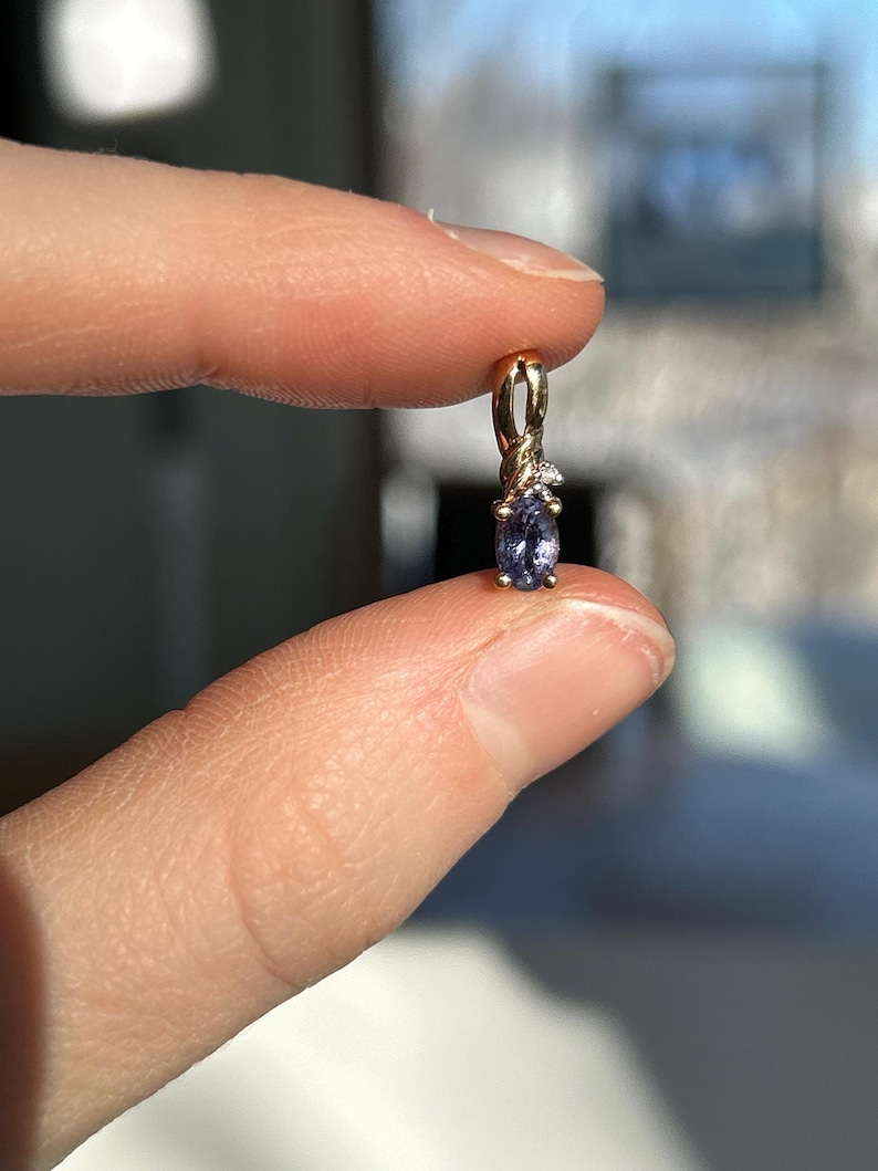Pode incluir: Um pingente de ouro com uma pequena pedra preciosa azul em forma oval. O pingente tem um design delicado com pequenas pedras de destaque transparentes. A joia &eacute; segurada entre dois dedos, com um fundo desfocado.